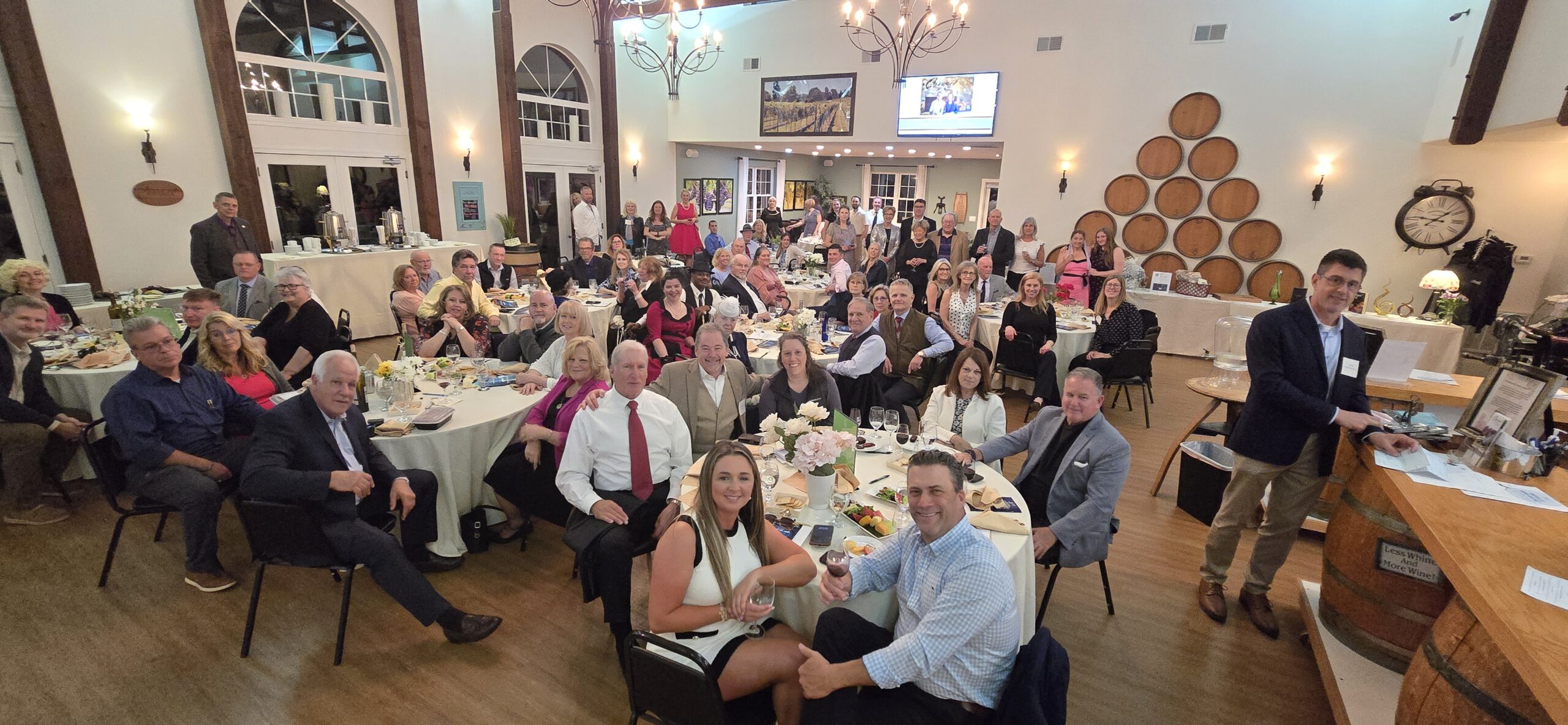 Large group of adults seated at round tables in a warmly lit event space with high ceilings and wine barrel decor. Most are dressed in business or semi-formal attire and smiling at the camera.